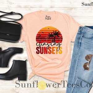 May include: A peach-colored t-shirt with a retro sunset graphic and the text "chasing sunsets". The graphic features palm trees and a red, orange, and yellow sunset. The t-shirt is paired with blue jeans, black boots, and a black purse.