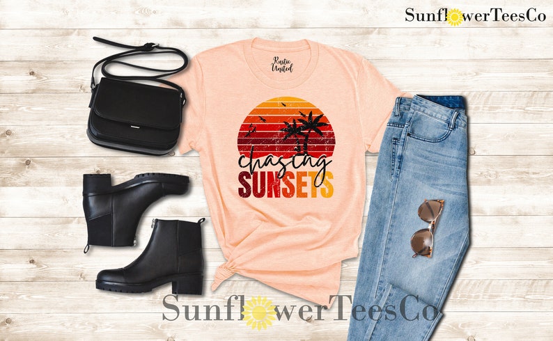 May include: A peach-colored t-shirt with a retro sunset graphic and the text "chasing sunsets". The graphic features palm trees and a red, orange, and yellow sunset. The t-shirt is paired with blue jeans, black boots, and a black purse.