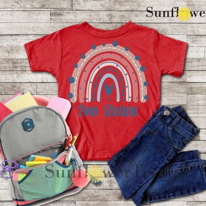 May include: Red t-shirt with a rainbow design and the text "Two Grade" on the front. The shirt is laid out on a wooden surface with a backpack, pencils, and jeans.