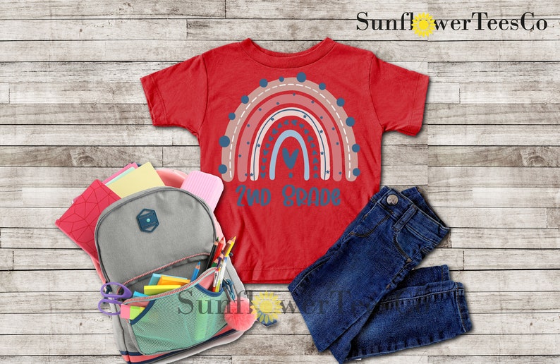 May include: Red t-shirt with a rainbow design and the text "Two Grade" on the front. The shirt is laid out on a wooden surface with a backpack, pencils, and jeans.