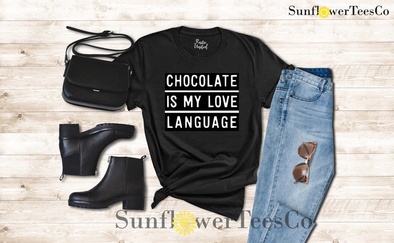 May include: Black t-shirt with the text "CHOCOLATE IS MY LOVE LANGUAGE" in a white rectangular box. The shirt is styled with black ankle boots, a black purse, and blue jeans with sunglasses.