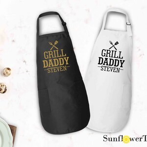 May include: Two black and white aprons with the text "Grill Daddy - Steven" and a crossed grill spatula and fork graphic. The aprons are available in black and white.