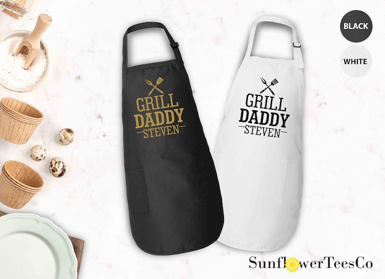 May include: Two black and white aprons with the text "Grill Daddy - Steven" and a crossed grill spatula and fork graphic. The aprons are available in black and white.