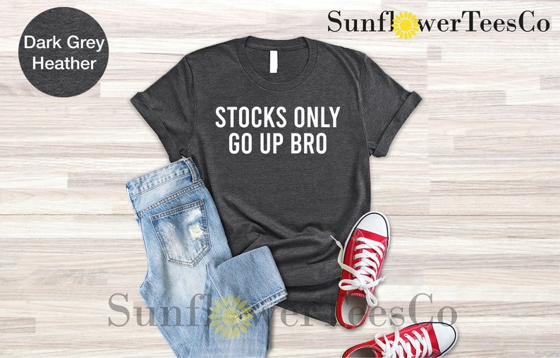 May include: Dark grey heather t-shirt with the text "STOCKS ONLY GO UP BRO" printed in white. The shirt is laid flat on a wooden surface with a pair of blue jeans and red sneakers.