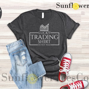 May include: A dark heather grey t-shirt with a white graphic that says "Lucky Trading Shirt - Do Not Wash". The graphic features a stylized chart with an upward arrow.