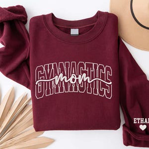 Op de afbeelding: Een bordeauxrode sweatshirt met de woorden "GYMNASTICS mom" in witte blokletters. De manchet heeft de naam "ETHAN" en een wit hart. Een bruine hoed is zichtbaar op de achtergrond.