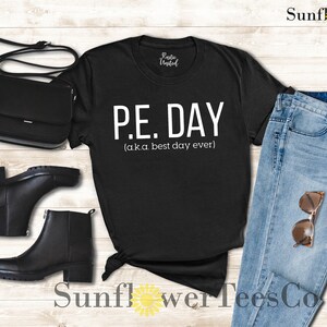 Op de afbeelding: Zwart T-shirt met witte tekst die "P.E. DAY (aka best day ever)" luidt. Het shirt ligt op een houten oppervlak met een paar zwarte laarzen, een zwarte tas en een blauwe jeans met een zonnebril.