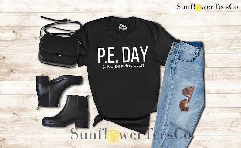 Op de afbeelding: Zwart T-shirt met witte tekst die "P.E. DAY (aka best day ever)" luidt. Het shirt ligt op een houten oppervlak met een paar zwarte laarzen, een zwarte tas en een blauwe jeans met een zonnebril.