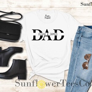 Op de afbeelding: Wit T-shirt met zwarte tekst die "DAD" zegt met "OLIVIA & JAMES" eronder. Het shirt ligt op een houten oppervlak met een paar zwarte laarzen, een zwarte tas en een blauwe jeans.
