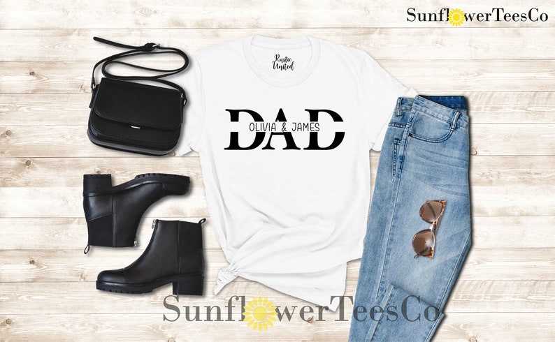 Op de afbeelding: Wit T-shirt met zwarte tekst die "DAD" zegt met "OLIVIA & JAMES" eronder. Het shirt ligt op een houten oppervlak met een paar zwarte laarzen, een zwarte tas en een blauwe jeans.