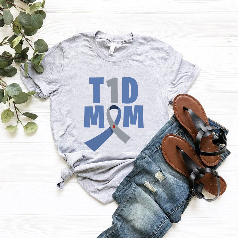 Pu&ograve; includere: Maglietta grigia con il testo "T1D MOM" e un nastro blu e grigio con un cuore rosso al centro.