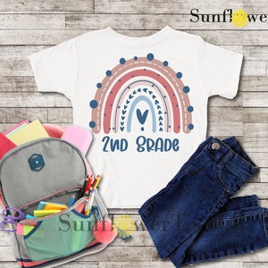 May include: White t-shirt with a rainbow design and the text "2nd Grade". The rainbow is made up of pink, red, blue, and white stripes with a heart in the center. The shirt is laid out on a wooden surface with a backpack, pencils, and jeans.
