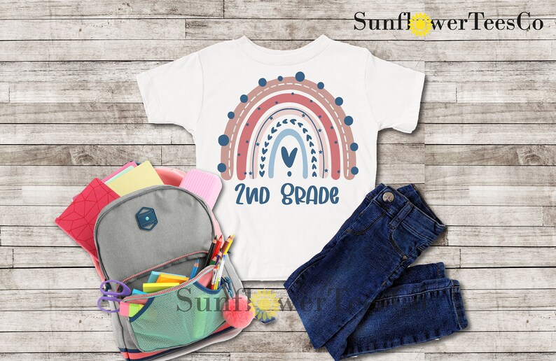 May include: White t-shirt with a rainbow design and the text "2nd Grade". The rainbow is made up of pink, red, blue, and white stripes with a heart in the center. The shirt is laid out on a wooden surface with a backpack, pencils, and jeans.
