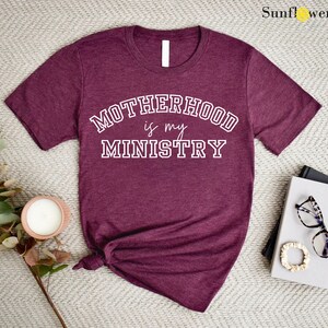 May include: A maroon t-shirt with the text "Motherhood is my Ministry" in white lettering. The text is in a collegiate font style.