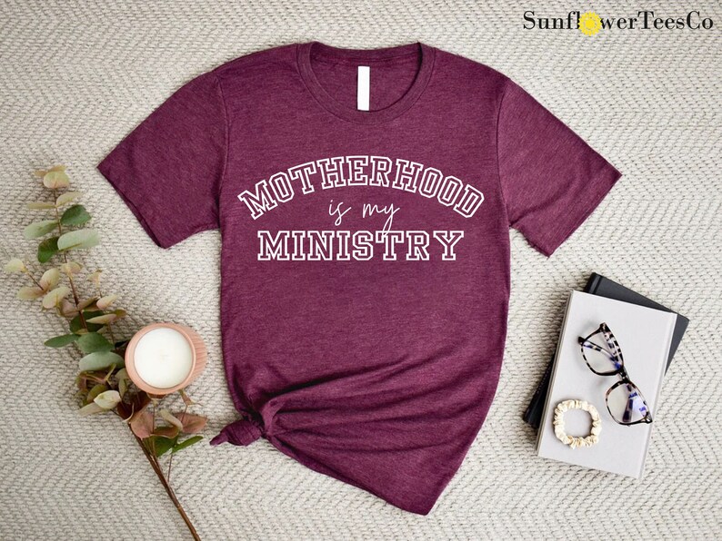 May include: A maroon t-shirt with the text "Motherhood is my Ministry" in white lettering. The text is in a collegiate font style.