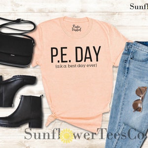 Op de afbeelding: Perzikkleurige T-shirt met zwarte tekst die "P.E. DAY (aka. best day ever)" luidt.