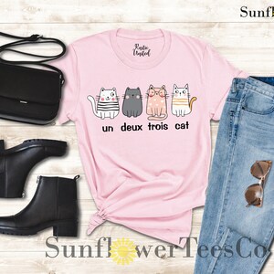 以下が含まれることがあります： ピンクのTシャツに、4匹の猫と「un deux trois cat」の文字がプリントされています。Tシャツは、青いジーンズ、黒いブーツ、黒いショルダーバッグと合わせています。