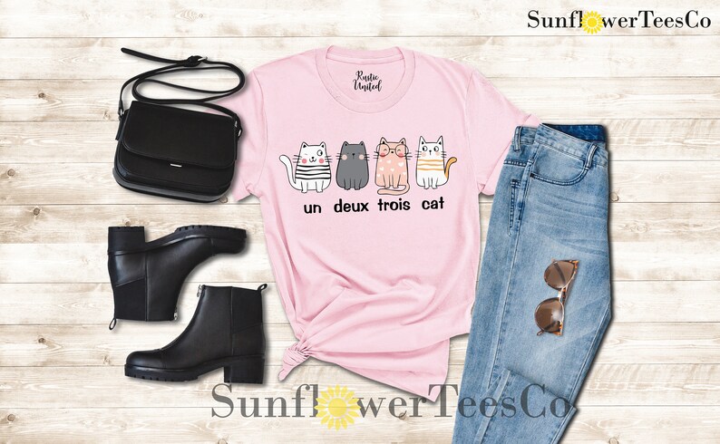以下が含まれることがあります： ピンクのTシャツに、4匹の猫と「un deux trois cat」の文字がプリントされています。Tシャツは、青いジーンズ、黒いブーツ、黒いショルダーバッグと合わせています。
