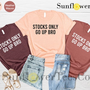 May include: Three t-shirts with the text "Stocks Only Go Up Bro" in white lettering on a maroon, peach, and rust colored background. The shirts are laid out on a wooden surface with denim shorts and sandals.