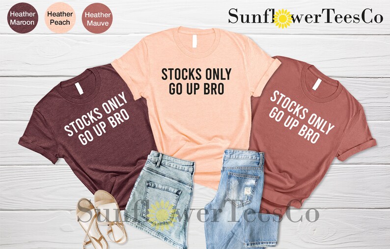 May include: Three t-shirts with the text "Stocks Only Go Up Bro" in white lettering on a maroon, peach, and rust colored background. The shirts are laid out on a wooden surface with denim shorts and sandals.