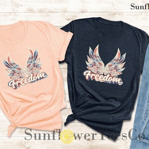 Puede incluir: Dos camisetas con un dise&ntilde;o de bandera estadounidense desgastado y la palabra "Freedom" en una fuente vintage. Las camisetas son de color melocot&oacute;n y azul marino.