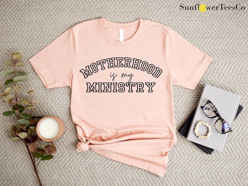 May include: A light pink t-shirt with the text "MOTHERHOOD is my MINISTRY" in a black collegiate font.