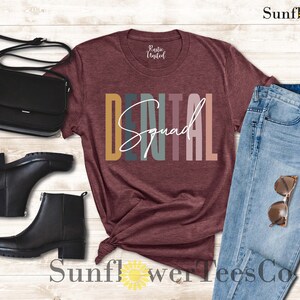 May include: A maroon t-shirt with the text "DENTAL Squad" in colorful letters. The shirt is styled with a pair of black boots, a black purse, and blue jeans.