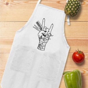 May include: White apron with two pockets and a black graphic of a hand making a rock and roll gesture, holding scissors and a comb. A pineapple, tomato, and green bell pepper are on a wooden surface.