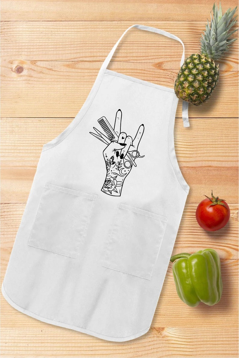 May include: White apron with two pockets and a black graphic of a hand making a rock and roll gesture, holding scissors and a comb. A pineapple, tomato, and green bell pepper are on a wooden surface.
