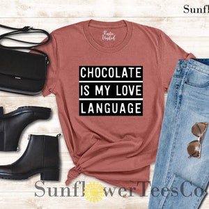 May include: A rust-colored t-shirt with the text "CHOCOLATE IS MY LOVE LANGUAGE" in a black and white design. The image also includes black ankle boots, a black purse, and blue jeans with sunglasses.