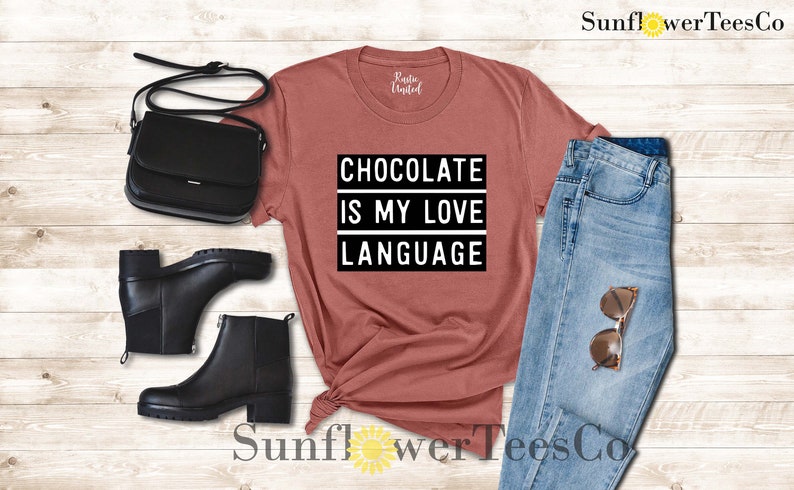 May include: A rust-colored t-shirt with the text "CHOCOLATE IS MY LOVE LANGUAGE" in a black and white design. The image also includes black ankle boots, a black purse, and blue jeans with sunglasses.