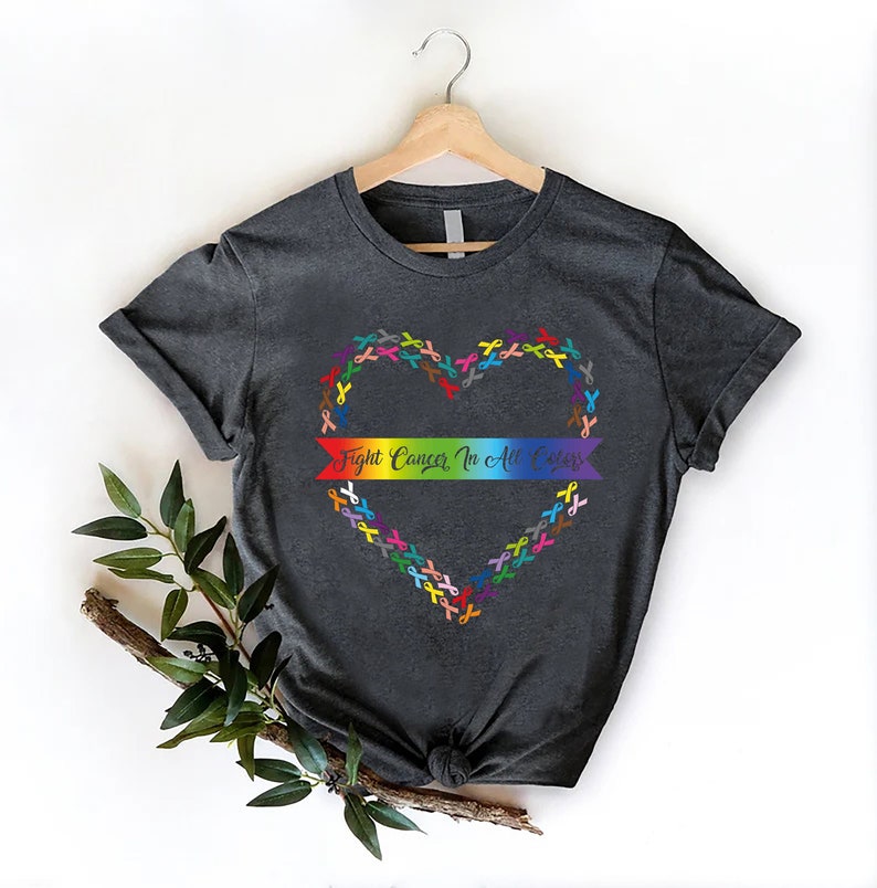May include: Dark gray t-shirt with a heart-shaped design made of colorful ribbons. A rainbow banner across the heart reads "Fight Cancer In All Colors". The shirt is displayed on a wooden hanger against a white background.