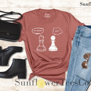 May include: A maroon t-shirt with a white graphic of two chess pieces. The white piece is thinking "Wait!" and the black piece is thinking "En Passant!"