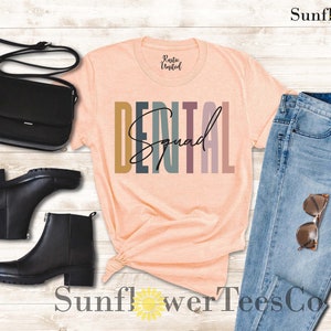 May include: A peach-colored t-shirt with the text "DENTAL Squad" in colorful, vertical stripes. The shirt is styled with a pair of black boots, a black purse, blue jeans, and sunglasses.