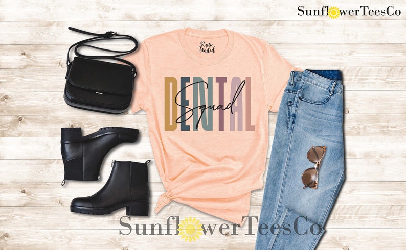 May include: A peach-colored t-shirt with the text "DENTAL Squad" in colorful, vertical stripes. The shirt is styled with a pair of black boots, a black purse, blue jeans, and sunglasses.