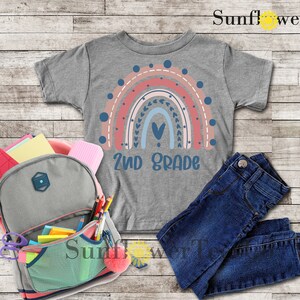 May include: A gray t-shirt with a rainbow design and the text "2nd Grade". The rainbow is made up of pink, red, blue, and white stripes with a heart in the center. The shirt is laid out on a wooden surface with a backpack, jeans, and school supplies.