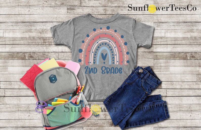 May include: A gray t-shirt with a rainbow design and the text "2nd Grade". The rainbow is made up of pink, red, blue, and white stripes with a heart in the center. The shirt is laid out on a wooden surface with a backpack, jeans, and school supplies.