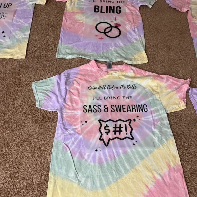 K&ouml;nnte beinhalten: Mehrere T-Shirts mit Batikmuster und aufgedruckten Slogans. Eines lautet "I'LL BRING THE BLING" mit Ringgrafiken. Ein anderes sagt "Raise Hell Before the Bells I'LL BRING THE SASS & SWEARING" mit einer Sprechblase.