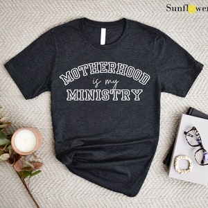 May include: A dark gray t-shirt with the text "MOTHERHOOD is my MINISTRY" in white lettering. The text is in a collegiate font.