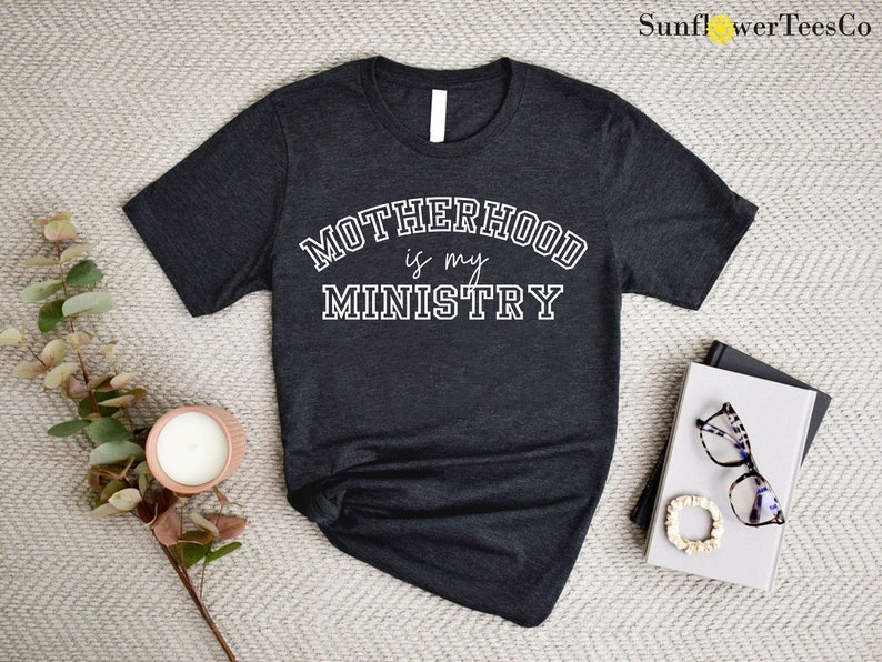 May include: A dark gray t-shirt with the text "MOTHERHOOD is my MINISTRY" in white lettering. The text is in a collegiate font.