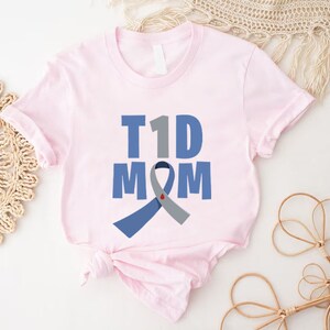 Pu&ograve; includere: Maglietta rosa con il testo "T1D MOM" e un nastro blu e grigio con un cuore rosso.