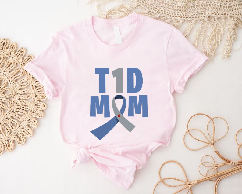 Pu&ograve; includere: Maglietta rosa con il testo "T1D MOM" e un nastro blu e grigio con un cuore rosso.