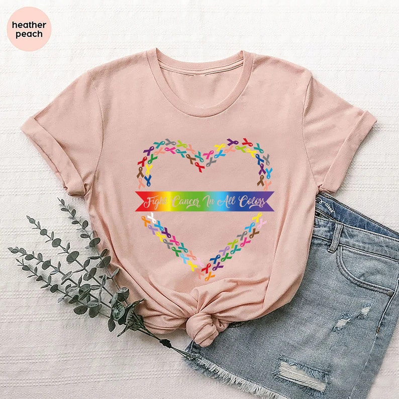 May include: A heather peach t-shirt featuring a heart made of colorful ribbons. The heart shape surrounds a rainbow banner with the text "Fight Cancer In All Colors". The shirt is paired with denim shorts.