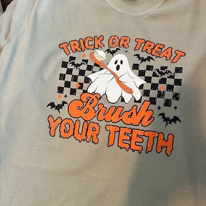 K&ouml;nnte beinhalten: Hellgr&uuml;nes T-Shirt mit einem Halloween-Motiv. Das Design zeigt den Text "Trick or Treat" und "Brush Your Teeth" in Orange, einen Cartoon-Geist mit einer Zahnb&uuml;rste, schwarze Flederm&auml;use und ein Schachbrettmuster. Ein lustiges, saisonales Design.