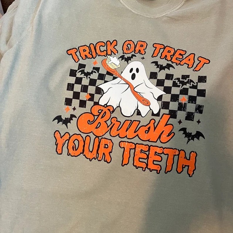 K&ouml;nnte beinhalten: Hellgr&uuml;nes T-Shirt mit einem Halloween-Motiv. Das Design zeigt den Text "Trick or Treat" und "Brush Your Teeth" in Orange, einen Cartoon-Geist mit einer Zahnb&uuml;rste, schwarze Flederm&auml;use und ein Schachbrettmuster. Ein lustiges, saisonales Design.