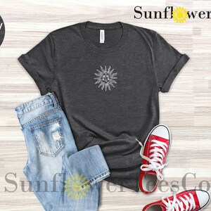May include: A dark heather grey t-shirt with a white sun graphic. The t-shirt is laid flat on a wooden surface with a pair of blue jeans and red sneakers.