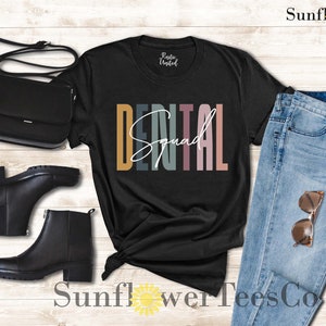 May include: Black t-shirt with the text "DENTAL Squad" in colorful letters. The shirt is displayed on a wooden background with a pair of black boots, a black purse, and blue jeans.