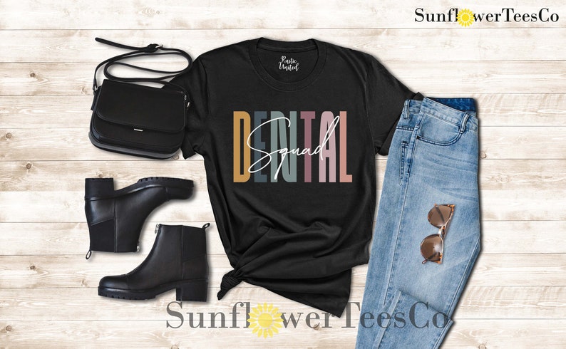May include: Black t-shirt with the text "DENTAL Squad" in colorful letters. The shirt is displayed on a wooden background with a pair of black boots, a black purse, and blue jeans.