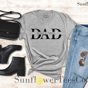 Op de afbeelding: Grijs T-shirt met zwarte tekst die "DAD Olivia & James" luidt. Het shirt wordt getoond op een houten oppervlak met een paar zwarte laarzen, een zwarte tas en een blauwe jeans.