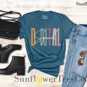 May include: A teal blue t-shirt with the text "Dental Squad" in colorful vertical stripes. The shirt is paired with blue jeans, black boots, and a black purse.